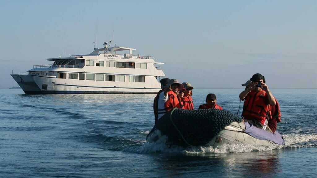 Toeristen aan boord van een pangaboot met de Millenium Galapagos-catamaran op de achtergrond